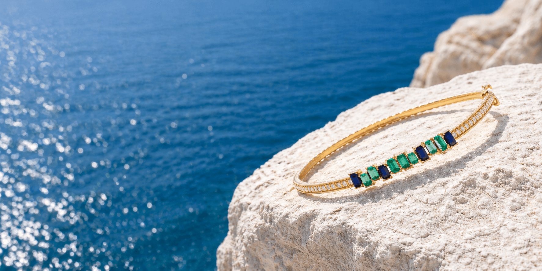 Gold bracelet set with sapphires and emeralds resting on a coastal rock above the sea